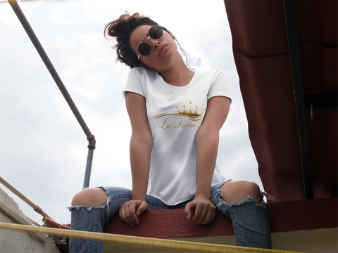 Woman sitting on a rooftop wearing a white shirt with the words in gold "La Reina" below a gold crown on top of a golden wave